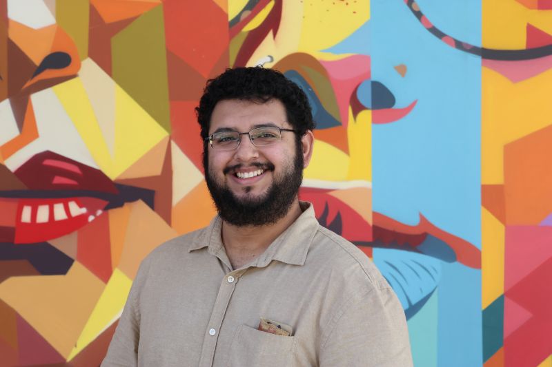 Portrait of Tutor Hector, smiling in front of colorful mural