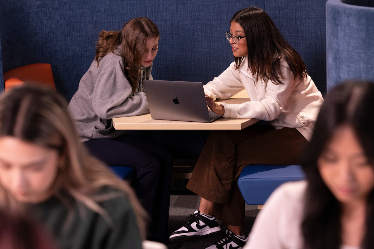 Two female students sit at a library table, looking at a laptop together.