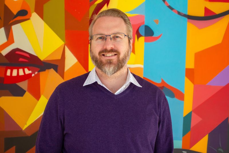 Headshot of Bruce Swanlund, smiling, wearing glasses and a purple sweater